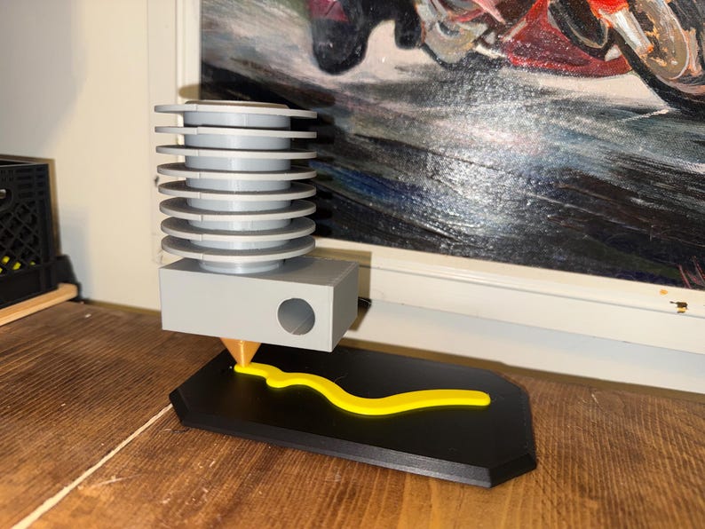 May include: A 3D printer head with a gray body and a yellow filament being extruded onto a black base. The printer head has a series of stacked, circular cooling fins. The base is a rectangular shape with a curved design.