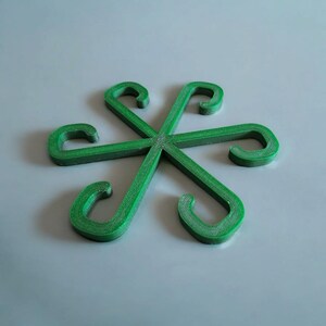 May include: A green, star-shaped object composed of six curved hooks. The hooks intersect at the center, forming a symmetrical design. The object is made of a solid, textured material and is set against a light gray background.