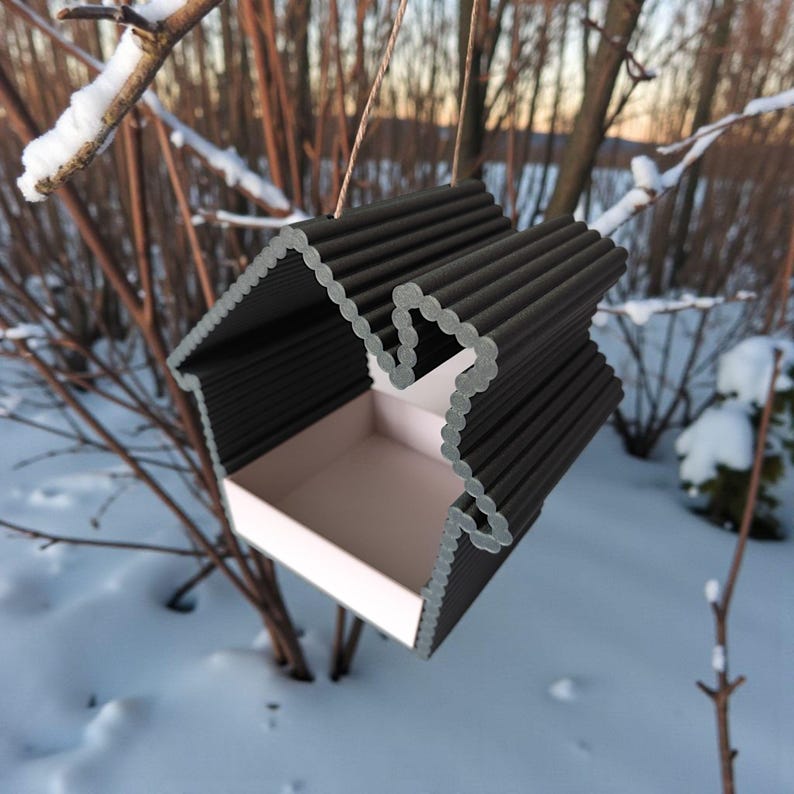 May include: A black and white bird feeder shaped like a house, hanging from a thin string. The feeder has a rectangular base and a corrugated roof. The background shows snow-covered branches and a snowy landscape.