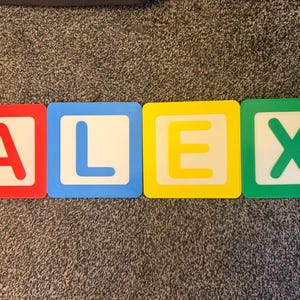 May include: Four colorful alphabet blocks spelling the name "ALEX". Each block has a white center with a letter in a different color: red, blue, yellow, and green. The blocks are on a gray carpet.