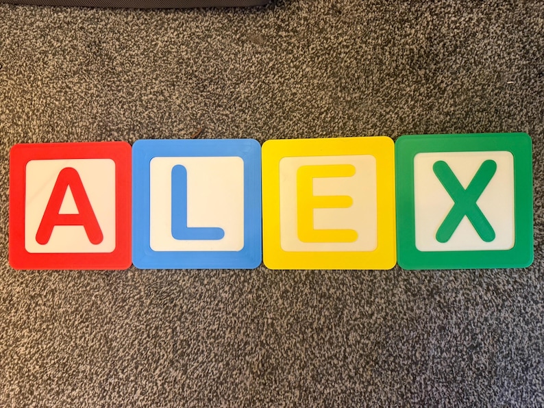May include: Four colorful alphabet blocks spelling the name "ALEX". Each block has a white center with a letter in a different color: red, blue, yellow, and green. The blocks are on a gray carpet.