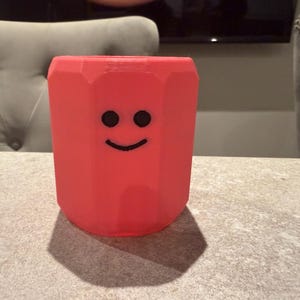 May include: A red pencil-shaped container with a smiling face. The container is a vibrant red color and has a hexagonal shape. The black eyes and smile are simple and add a playful touch to the design. The container is sitting on a table.