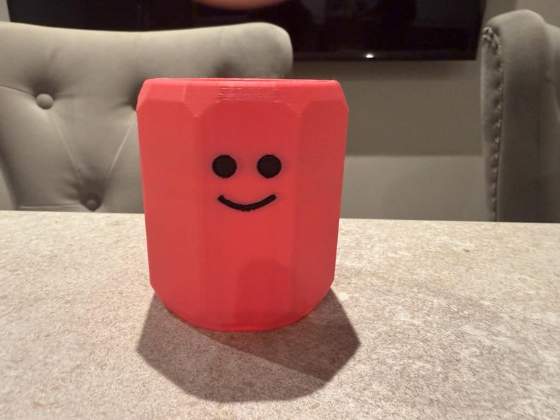 May include: A red pencil-shaped container with a smiling face. The container is a vibrant red color and has a hexagonal shape. The black eyes and smile are simple and add a playful touch to the design. The container is sitting on a table.