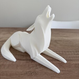 May include: A white, stylized sculpture of a howling wolf. The wolf is depicted in a seated position with its head raised and mouth open. The sculpture is made of a smooth, solid material and sits on a wooden surface.