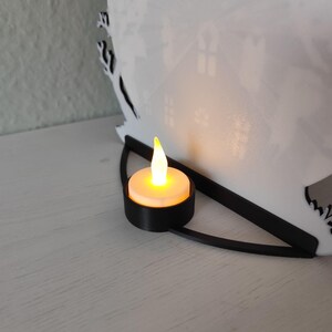 Pu&ograve; includere: Una lanterna di carta bianca tagliata al laser con un design a silhouette di casa. Una piccola candela a batteria &egrave; posta davanti alla lanterna, emettendo un bagliore caldo. La lanterna ha una base nera curva.