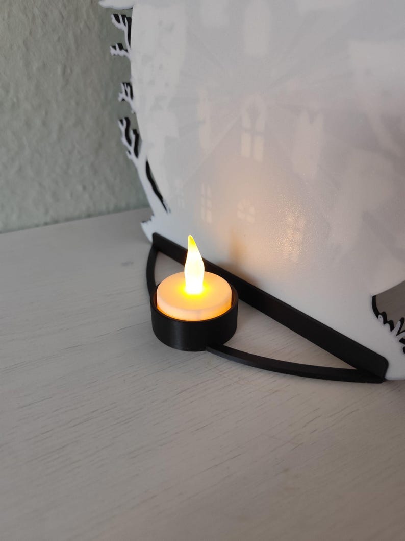 Pu&ograve; includere: Una lanterna di carta bianca tagliata al laser con un design a silhouette di casa. Una piccola candela a batteria &egrave; posta davanti alla lanterna, emettendo un bagliore caldo. La lanterna ha una base nera curva.