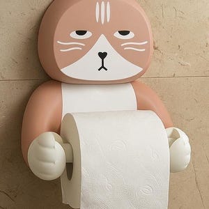 May include: A novelty cat-shaped toilet paper holder in peach and white. The holder is designed to look like a cat's head and arms, holding a roll of white toilet paper. The cat has a grumpy expression.