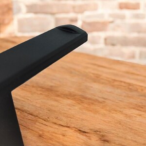 May include: Close-up of a black, angled furniture leg with a small indentation on the top edge. The leg is positioned over a wooden surface with visible grain. A brick wall is in the background.