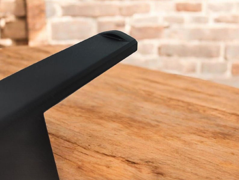May include: Close-up of a black, angled furniture leg with a small indentation on the top edge. The leg is positioned over a wooden surface with visible grain. A brick wall is in the background.