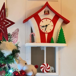 May include: A red and white Christmas clock with a candy cane and green tree decorations. The clock has a white face with black hands and a small figure in the top window. A shelf below holds a candy and a plush toy.