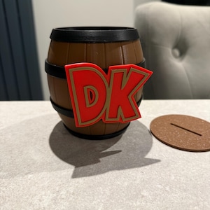 May include: A brown barrel-shaped container with black bands and a red and gold "DK" logo. A brown, circular lid with a slot sits beside the container. The container is on a light gray surface.