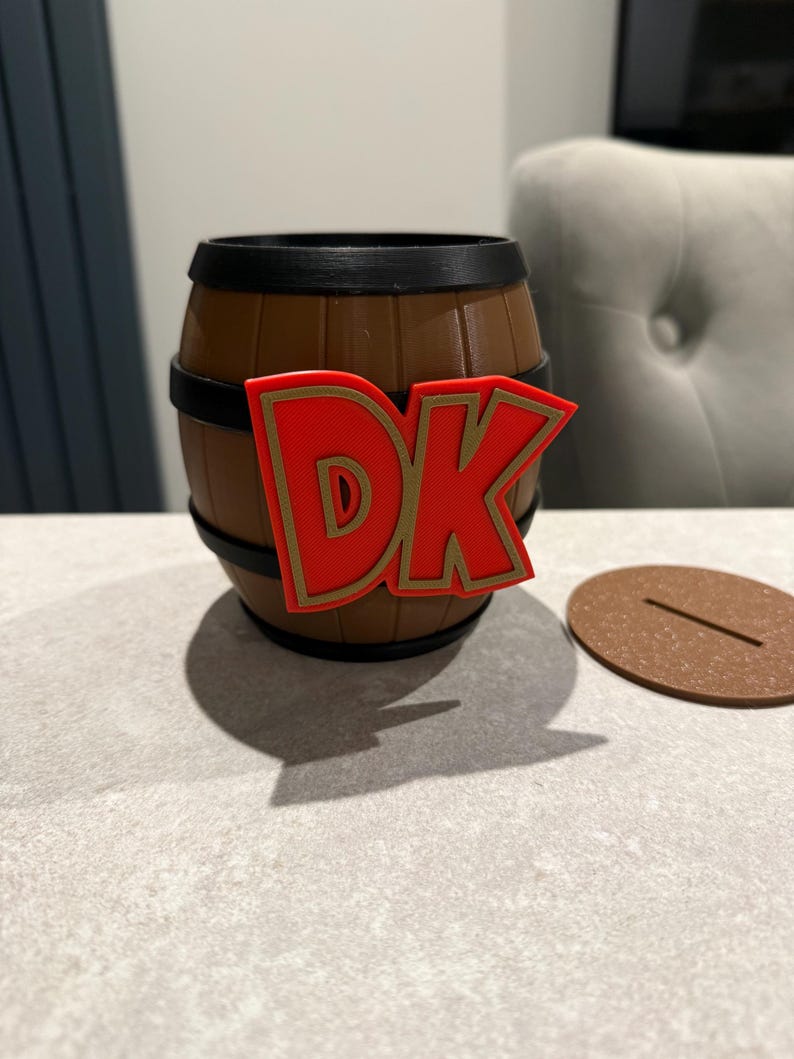 May include: A brown barrel-shaped container with black bands and a red and gold "DK" logo. A brown, circular lid with a slot sits beside the container. The container is on a light gray surface.