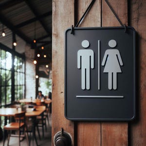 Puede incluir: Un letrero de ba&ntilde;o negro con figuras blancas de hombre y mujer. El letrero rectangular con esquinas redondeadas cuelga de una puerta de madera. El fondo muestra el interior de un restaurante.