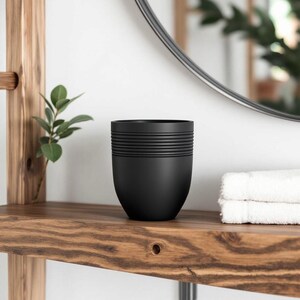 May include: A black, modern tumbler with horizontal ridged detailing near the top. The tumbler sits on a wooden shelf with a small plant and a stack of white towels. A round mirror is in the background.