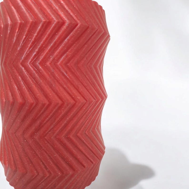 May include: A red, cylindrical object with a textured, zigzag pattern. The object has a slightly glittery finish and is set against a white background. The design creates a visually interesting, geometric effect.