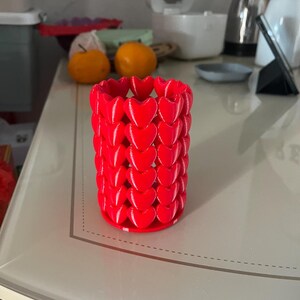 Puede incluir: Un recipiente cil&iacute;ndrico rojo brillante, adornado con un patr&oacute;n de corazones repetidos. El recipiente est&aacute; hecho de un material s&oacute;lido y se asienta sobre una base plana. El dise&ntilde;o es moderno y llamativo, adecuado para diversos usos decorativos.
