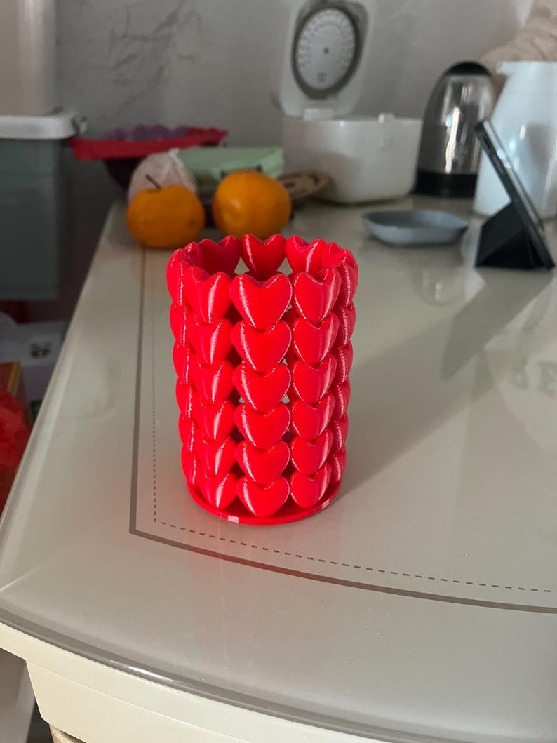 Puede incluir: Un recipiente cil&iacute;ndrico rojo brillante, adornado con un patr&oacute;n de corazones repetidos. El recipiente est&aacute; hecho de un material s&oacute;lido y se asienta sobre una base plana. El dise&ntilde;o es moderno y llamativo, adecuado para diversos usos decorativos.