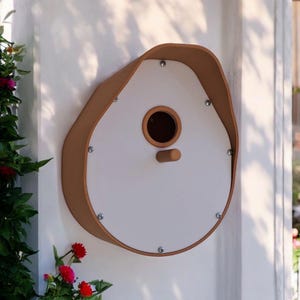 May include: A white birdhouse with a brown leather-like trim. The birdhouse has a round opening for birds to enter. The birdhouse is mounted on a white wall.