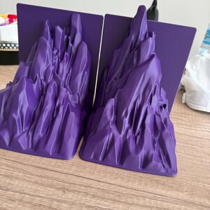 May include: Two purple bookends with a mountain range design. Each bookend has a flat, rectangular base and a sculpted mountain range in a vibrant purple color. The bookends are made of a smooth material and are designed to hold books upright.