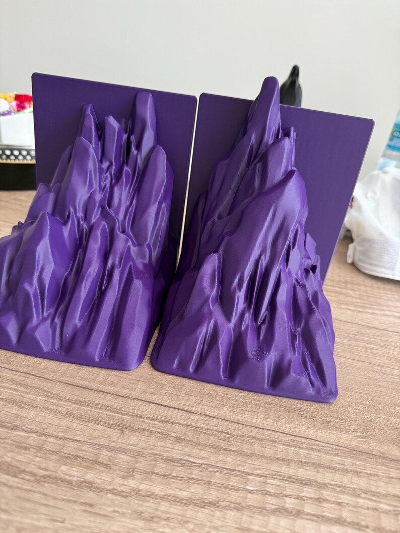 May include: Two purple bookends with a mountain range design. Each bookend has a flat, rectangular base and a sculpted mountain range in a vibrant purple color. The bookends are made of a smooth material and are designed to hold books upright.