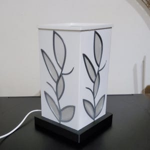 May include: A white, rectangular table lamp with a black base. The lamp features a leaf design in grey and black on each side. The lamp is plugged into a wall outlet.