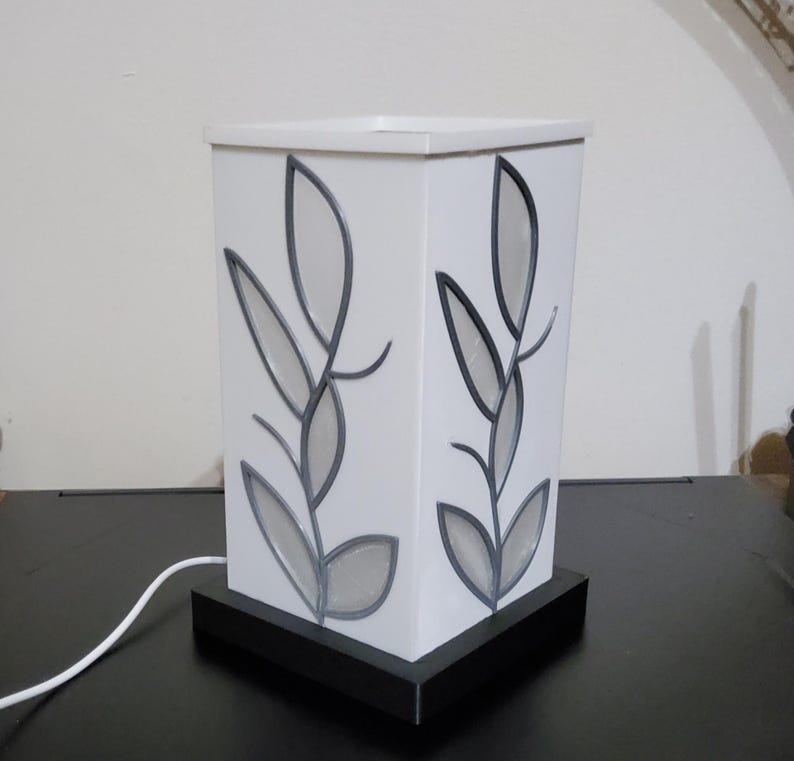 May include: A white, rectangular table lamp with a black base. The lamp features a leaf design in grey and black on each side. The lamp is plugged into a wall outlet.