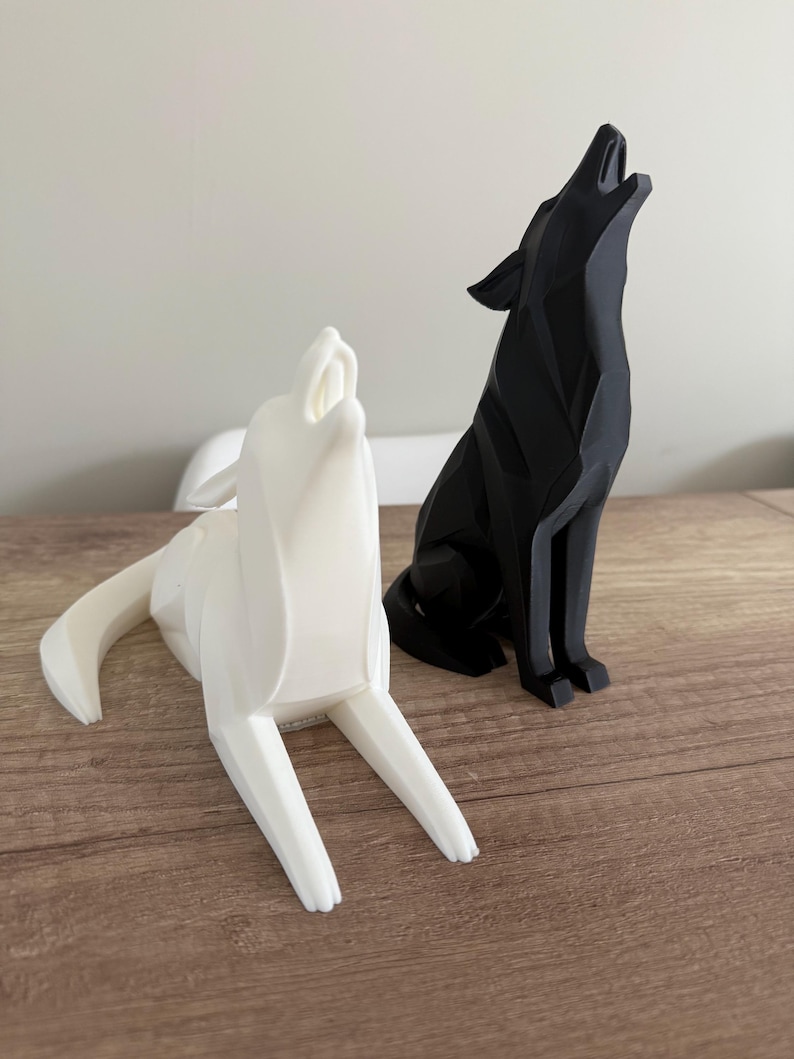 May include: Two geometric wolf sculptures. One is white and the other is black. Both are in a howling pose, with their heads tilted back and mouths open. The sculptures are placed on a wooden surface.