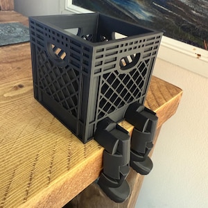 May include: A black plastic crate with a grid pattern, resting on a wooden surface. The crate has two handle openings and is supported by two black, leg-like attachments, giving the appearance of sitting.
