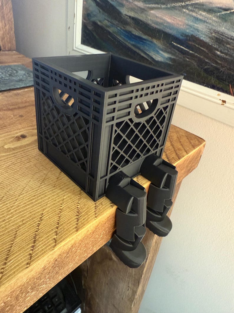May include: A black plastic crate with a grid pattern, resting on a wooden surface. The crate has two handle openings and is supported by two black, leg-like attachments, giving the appearance of sitting.