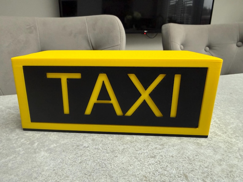 May include: A yellow taxi cab roof sign with the word "TAXI" in bold, white letters against a black background. The sign is rectangular and sits on a light gray surface.