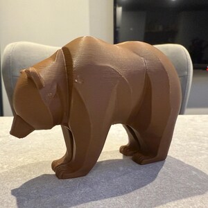 May include: A brown bear figurine with a geometric design. The bear is standing on all fours, with its head slightly lowered. The figurine is made of a solid material and has a textured surface. The bear is a decorative item.