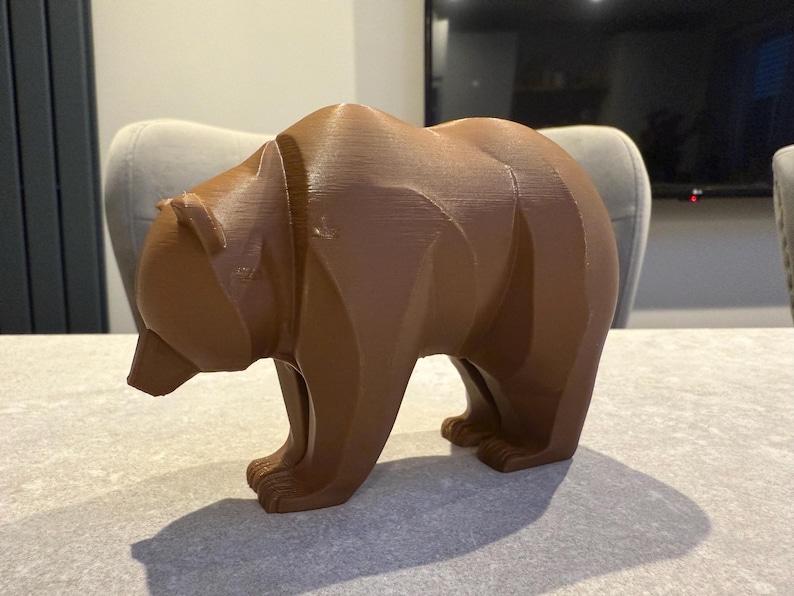 May include: A brown bear figurine with a geometric design. The bear is standing on all fours, with its head slightly lowered. The figurine is made of a solid material and has a textured surface. The bear is a decorative item.