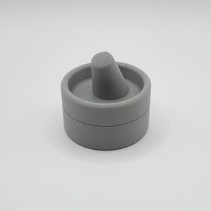 Pode incluir: Uma caixa de anel cinza, impressa em 3D, com um suporte c&ocirc;nico. A caixa &eacute; redonda com tampa e base. O suporte do anel &eacute; centralizado dentro da caixa e tem um topo canelado.