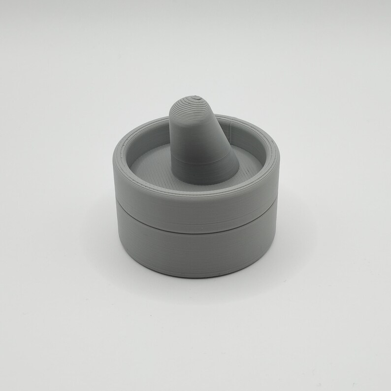 Pode incluir: Uma caixa de anel cinza, impressa em 3D, com um suporte c&ocirc;nico. A caixa &eacute; redonda com tampa e base. O suporte do anel &eacute; centralizado dentro da caixa e tem um topo canelado.