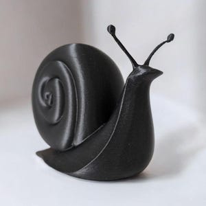 May include: A black, 3D-printed snail figurine. The snail has a spiraled shell and two antennae with small, round tips. The figurine is a solid, matte black color and sits on a white surface.