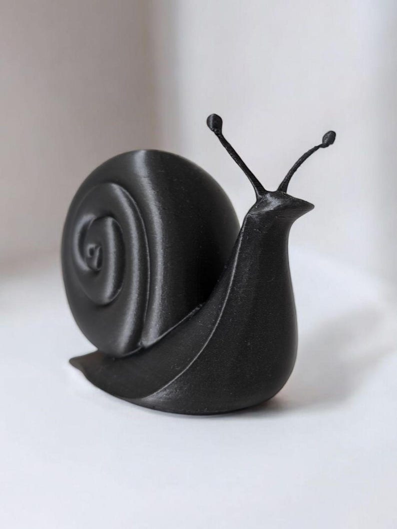 May include: A black, 3D-printed snail figurine. The snail has a spiraled shell and two antennae with small, round tips. The figurine is a solid, matte black color and sits on a white surface.