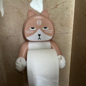 May include: A novelty toilet paper holder in the shape of a cat's face. The holder is peach and white, with a bored expression. The toilet paper roll is white and visible. The holder is mounted on a beige tiled wall.