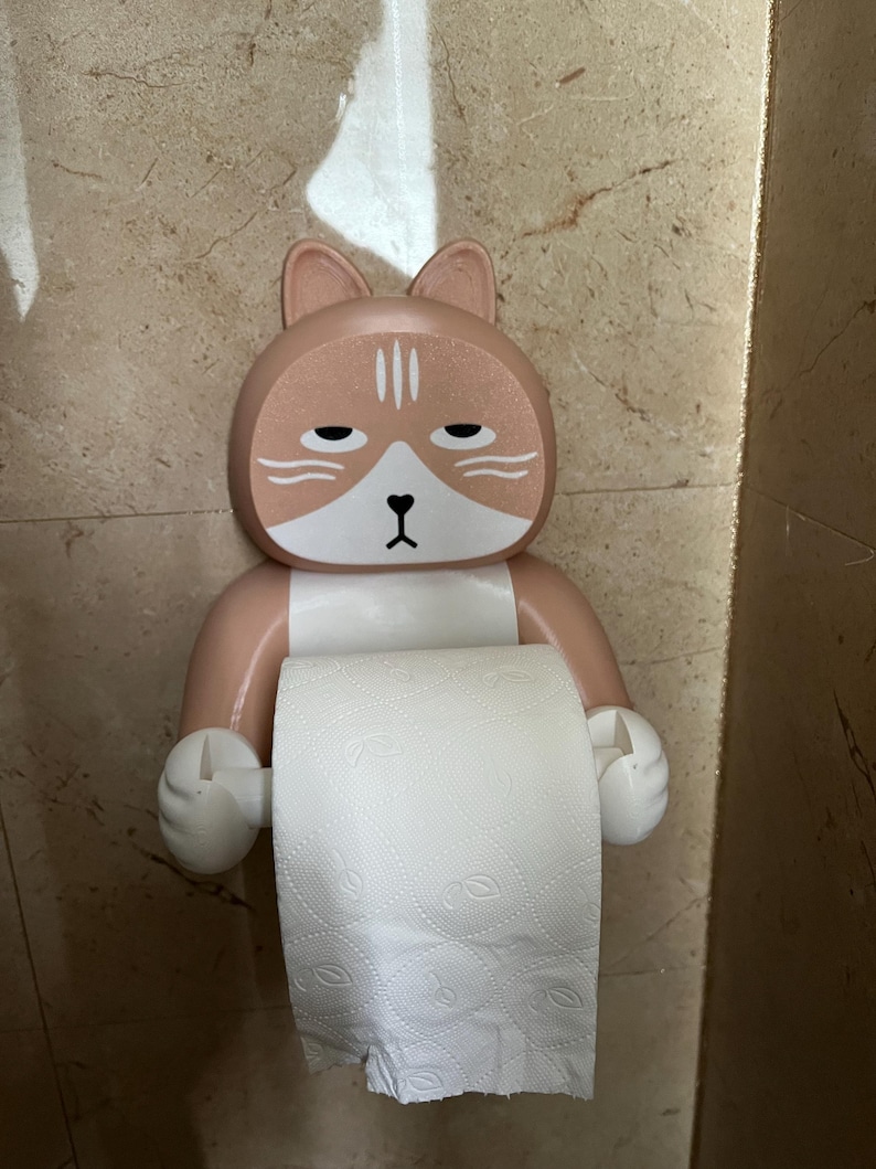 May include: A novelty toilet paper holder in the shape of a cat's face. The holder is peach and white, with a bored expression. The toilet paper roll is white and visible. The holder is mounted on a beige tiled wall.