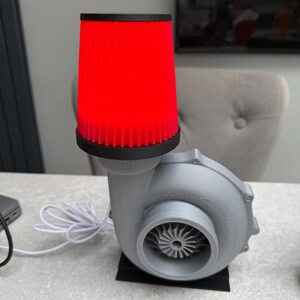 May include: A gray, turbine-shaped lamp with a bright red cylindrical shade. The lamp has a black base and a white power cord. The shade is made of a ribbed material. The lamp is designed to resemble an automotive turbocharger.