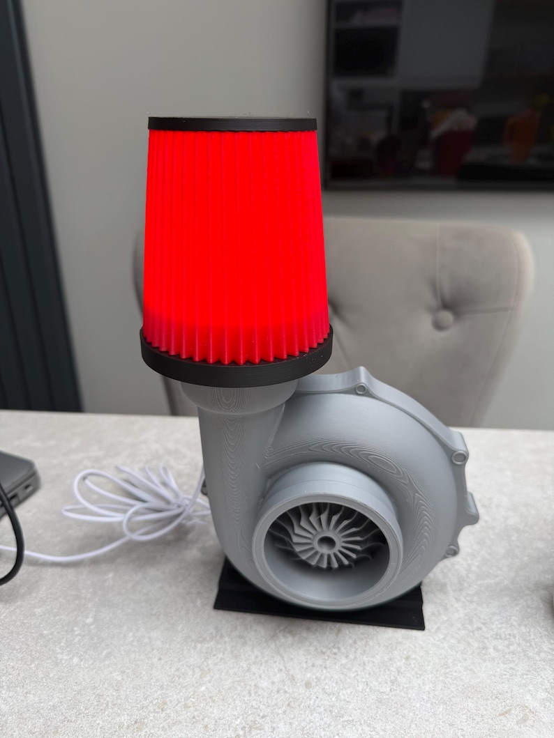 May include: A gray, turbine-shaped lamp with a bright red cylindrical shade. The lamp has a black base and a white power cord. The shade is made of a ribbed material. The lamp is designed to resemble an automotive turbocharger.