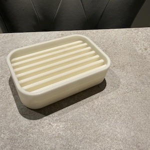 May include: A rectangular, off-white soap dish with a slatted top. The dish is made of a solid material and sits on a gray surface. The design allows for water drainage, keeping soap dry and lasting longer.