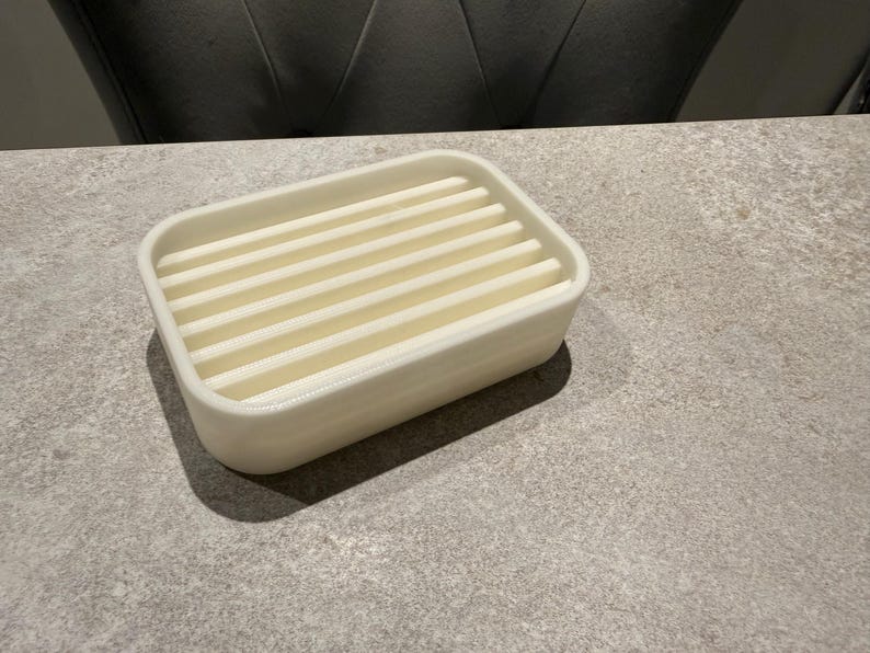 May include: A rectangular, off-white soap dish with a slatted top. The dish is made of a solid material and sits on a gray surface. The design allows for water drainage, keeping soap dry and lasting longer.
