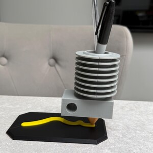 May include: A 3D-printed pen holder with a unique design. The holder features a gray cylindrical section with stacked rings, a gray rectangular base, and a black base with a yellow, wavy line. Two pens are inserted into the top. The design is modern and functional.