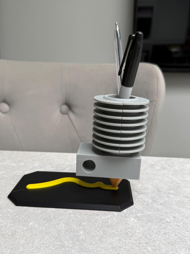 May include: A 3D-printed pen holder with a unique design. The holder features a gray cylindrical section with stacked rings, a gray rectangular base, and a black base with a yellow, wavy line. Two pens are inserted into the top. The design is modern and functional.