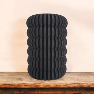 May include: A black, cylindrical vase with a textured, ribbed design. The vase is made of a dark material and sits on a wooden surface. The vase has a unique, modern aesthetic.