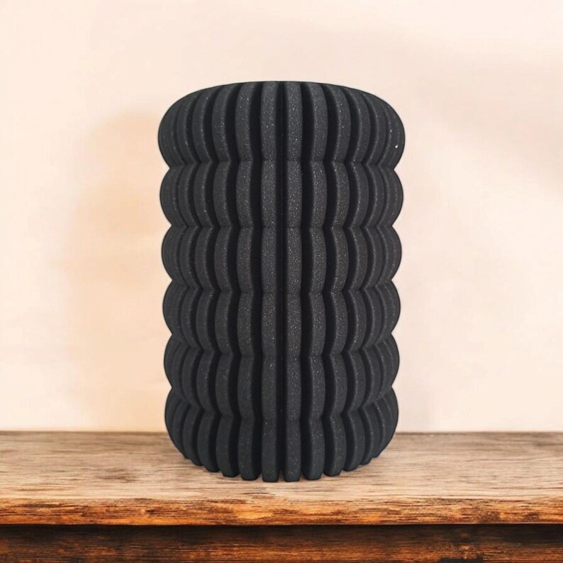 May include: A black, cylindrical vase with a textured, ribbed design. The vase is made of a dark material and sits on a wooden surface. The vase has a unique, modern aesthetic.