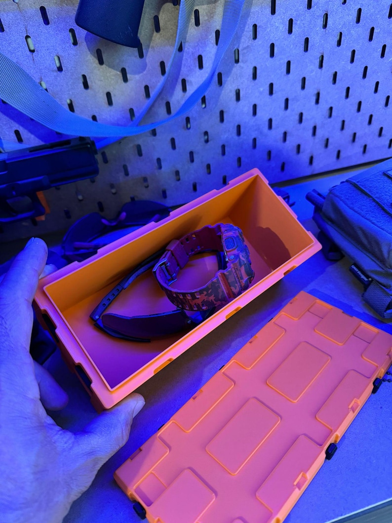 May include: An open orange storage box containing a camouflage watch and sunglasses. The box is rectangular with a lid and is sitting on a surface. The background includes a pegboard and other items.