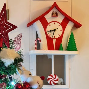 May include: A red and white Christmas clock with a candy cane and green tree decorations. The clock has a white face with black hands and a small figure in the top window. A shelf below holds a candy and a plush toy.