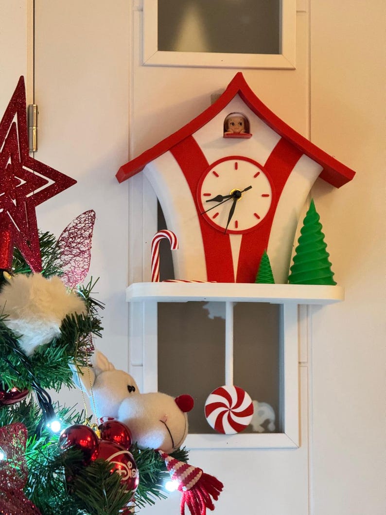 May include: A red and white Christmas clock with a candy cane and green tree decorations. The clock has a white face with black hands and a small figure in the top window. A shelf below holds a candy and a plush toy.