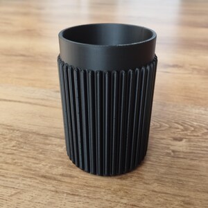 May include: A black, cylindrical cup with a ribbed, textured exterior. The cup has a smooth, rounded top and a textured, ridged lower section. The cup is made of a solid, opaque material.